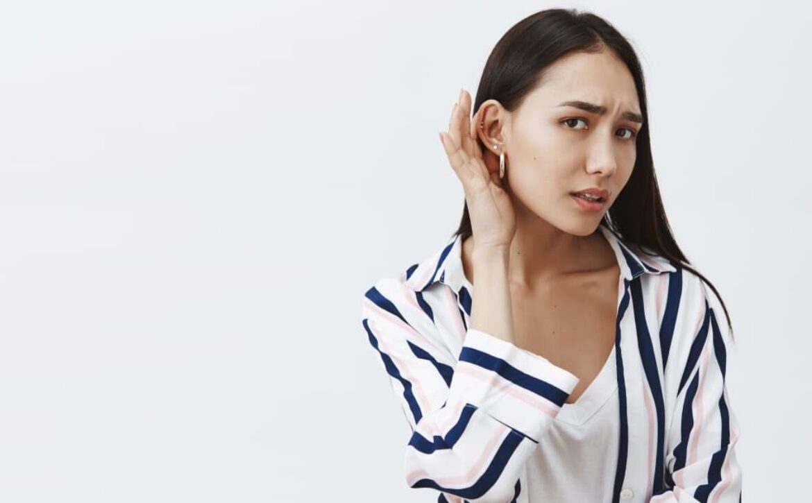 Studio shot of good-looking gloomy girlfriend with long dark hair, leaning towards camera with one ear and gazing at camera, eavesdropping and whispering, hearing rumor, feeling nervous