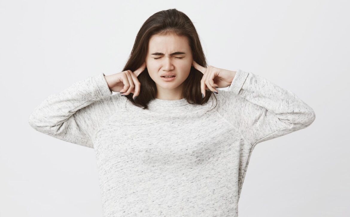 Studio shot of cute brunette student, epxressing stress and pressure, holding fingers in ears not to hear something and closing her eyes, over white background. Girl can not focus at all