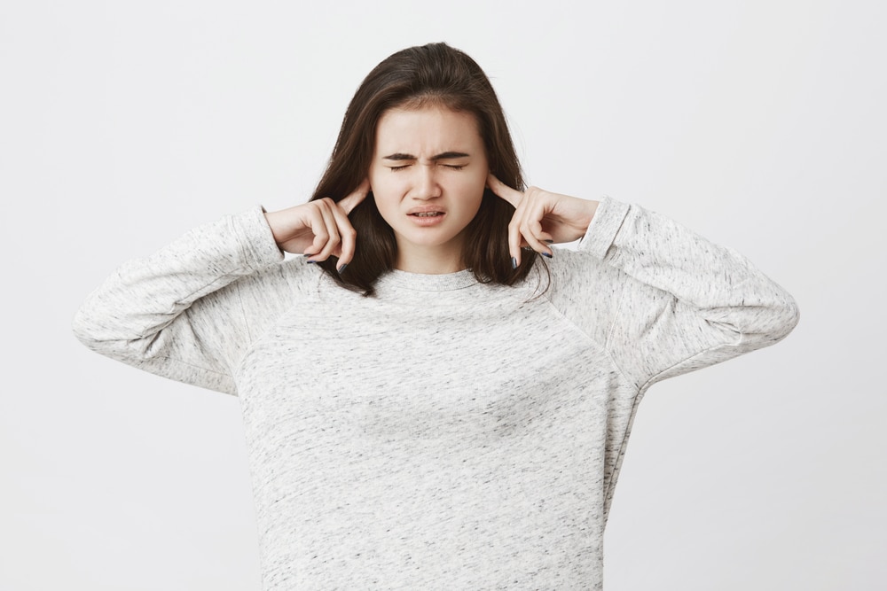 Studio shot of cute brunette student, epxressing stress and pressure, holding fingers in ears not to hear something and closing her eyes, over white background. Girl can not focus at all
